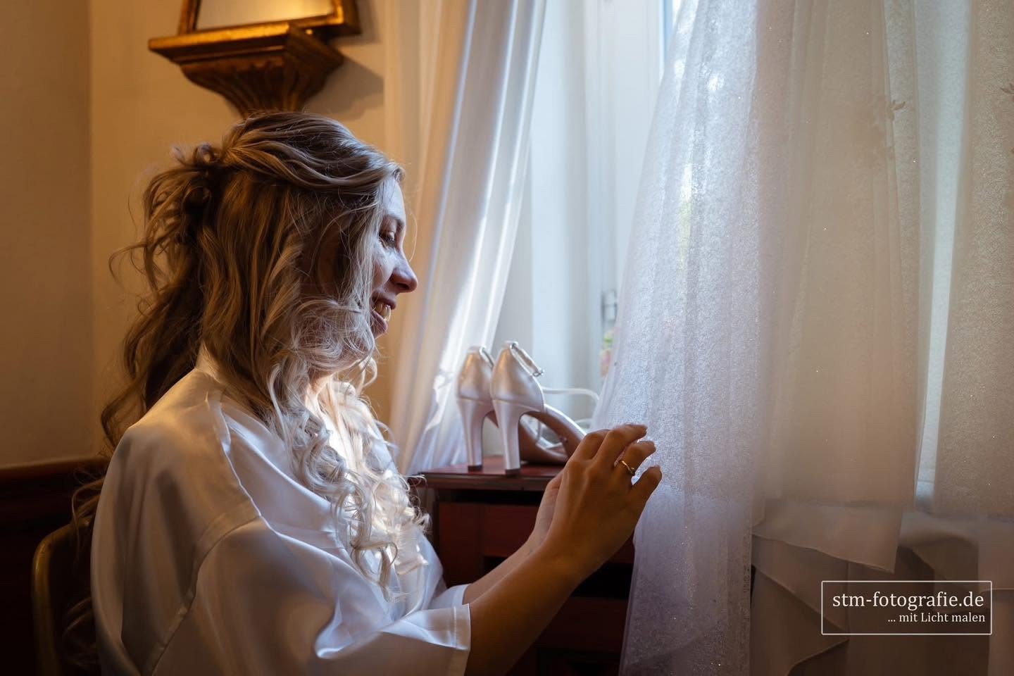 Braut im Morgenmantel schaut während der Hochzeitsvorbereitungen lächelnd zum Fenster